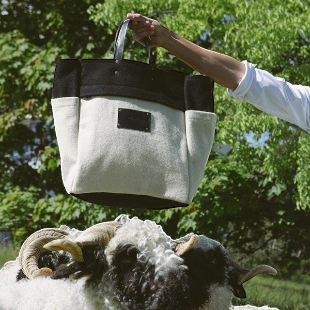 Baabuk Blacknose tote bag in cream and black wool with leather handles, held above a Valais Blacknose sheep.