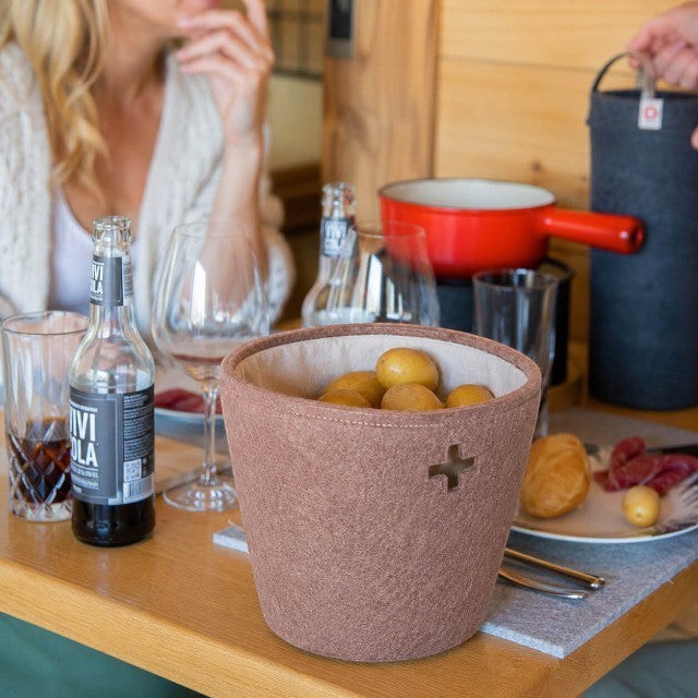 Baabuk small brown felted wool basket with Swiss cross cutout, holding potatoes on a dining table.