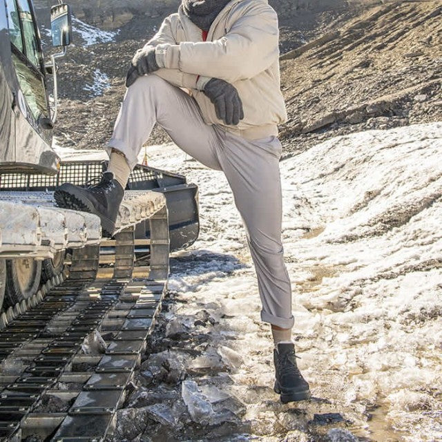 Man wearing Baabuk Ice Wooler black wool boots standing on snowy terrain next to a snow groomer