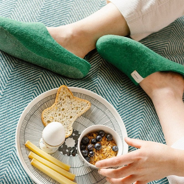 Baabuk Kanga lush green wool slippers worn by a person sitting on a patterned rug with a breakfast plate