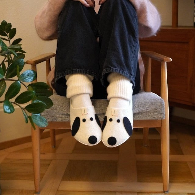 Baabuk Kanga Woof slippers in white wool with black dog-inspired features worn by a person sitting in a chair.