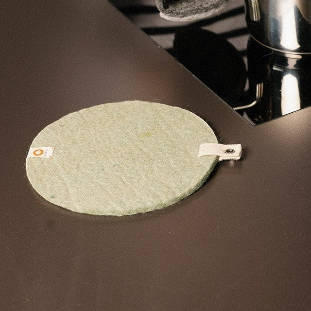 Baabuk round khaki felted wool trivet with a leather strap on a dark kitchen countertop.