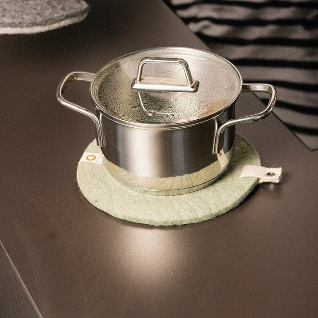 Baabuk khaki felted wool trivet hot plate with a leather strap under a stainless steel pot on a dark table