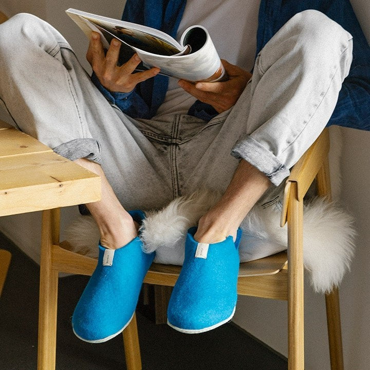 Baabuk Lisa slippers in azur blue wool worn by a person sitting on a wooden chair reading a magazine