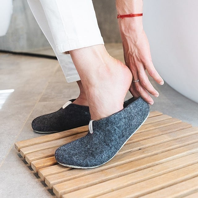 Baabuk Mel dark grey wool slippers with natural crepe rubber soles being worn on a wooden bath mat